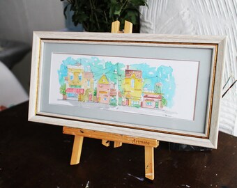 May include: A watercolor painting of a row of colorful houses with a blue sky and white clouds in the background. The painting is framed in a white frame and is displayed on a wooden easel.