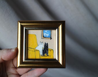 May include: A miniature painting in a gold frame showing a white and a black cat on a yellow sofa. The background is beige with a blue window. The artwork is a small, framed piece.