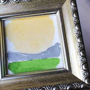 May include: A small framed watercolor painting of a landscape with a yellow sun, gray hills, and green grass.