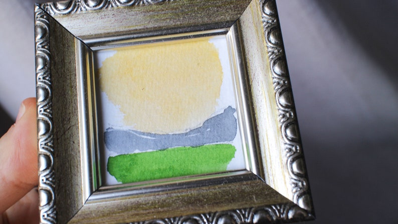 May include: A small framed watercolor painting of a landscape with a yellow sun, gray hills, and green grass.