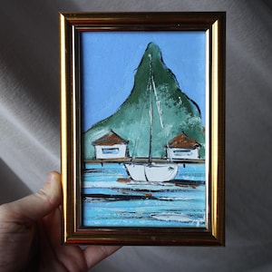 May include: A framed painting of a sailboat in front of a green island with two small white houses. The water is blue and the sky is light blue.