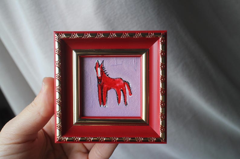 A small, square framed artwork featuring a red horse on a lavender background. The frame is red with gold trim. The artwork is held in a hand.