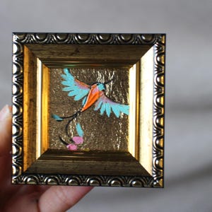 May include: A small framed painting of a blue and orange bird with a long tail, set against a gold background.