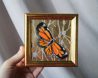May include: A framed painting of a monarch butterfly with orange and black wings on a gold background with white accents.