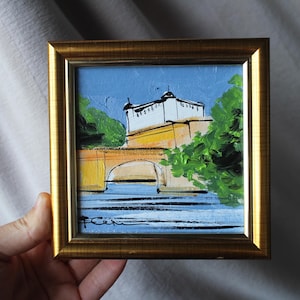 A small framed painting depicting a bridge and building with a blue sky background. The artwork features a gold frame and is held by a hand. The painting uses yellow, white, green, and blue colors.