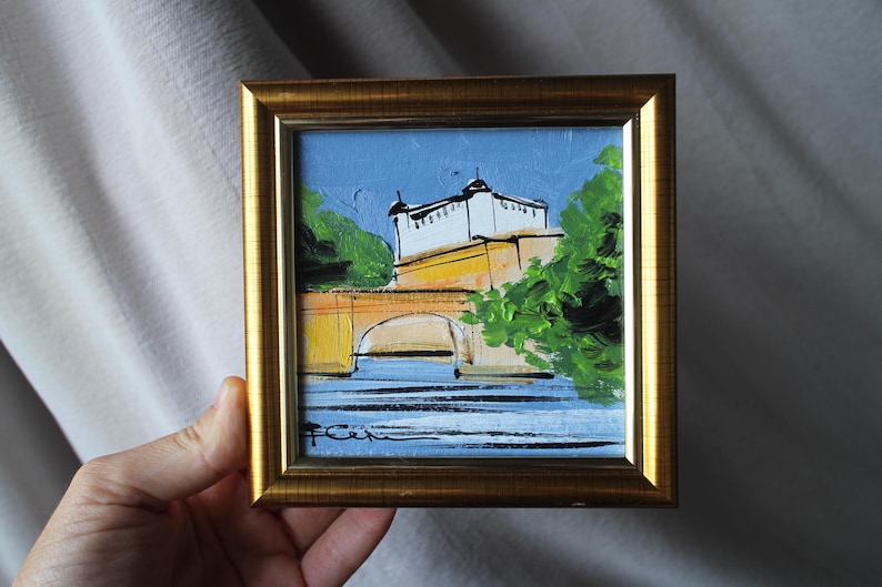 A small framed painting depicting a bridge and building with a blue sky background. The artwork features a gold frame and is held by a hand. The painting uses yellow, white, green, and blue colors.