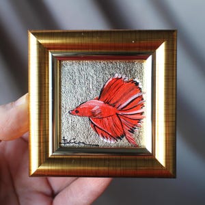 May include: Miniature framed painting of a red betta fish with a flowing tail. The artwork is set against a silver background and framed in a gold-colored frame. The painting is held in a hand.