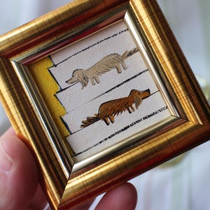 May include: A framed painting of two dogs walking up a set of stairs. The dogs are painted in a simple, cartoonish style. The top dog is light brown and the bottom dog is brown. The stairs are painted in yellow and white.