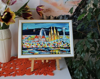 May include: A colorful painting of a cityscape with buildings, a lighthouse, and a cable car. The painting is framed in white with a gold trim and is displayed on a wooden easel. The painting is on a white surface with a red and orange patterned mat.