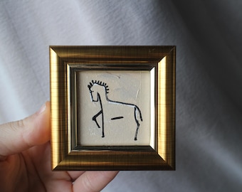 May include: Small framed artwork featuring a minimalist black and white horse illustration. The artwork is set within a gold-colored square frame. The horse is depicted with simple lines and shapes, creating a modern, artistic aesthetic.