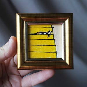 May include: A framed miniature painting depicts a black and white cat lounging on yellow stairs. The artwork is set within a gold-colored frame. The cat is painted with fine detail, adding a touch of whimsy to the piece.