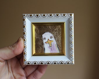 May include: A framed painting of a white bird with a yellow beak and pink accents on a gold background.