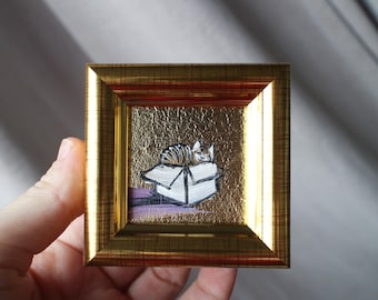 May include: A small, gold-framed painting of a cat curled up inside a cardboard box. The cat is brown and white, and the box is white with a brown top. The background is a gold-colored textured surface.
