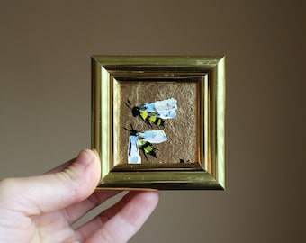 May include: A small framed painting of two bees with yellow and black stripes on a gold background.