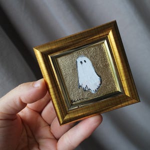 K&ouml;nnte beinhalten: Ein kleines, quadratisches, gerahmtes Gem&auml;lde mit einem wei&szlig;en Geist mit schwarzen Augen auf goldenem Hintergrund. Der Rahmen ist goldfarben. Das Kunstwerk wird in einer Hand gehalten.