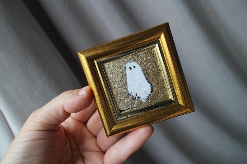 K&ouml;nnte beinhalten: Ein kleines, quadratisches, gerahmtes Gem&auml;lde mit einem wei&szlig;en Geist mit schwarzen Augen auf goldenem Hintergrund. Der Rahmen ist goldfarben. Das Kunstwerk wird in einer Hand gehalten.