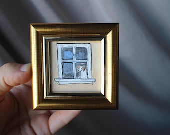 Może przedstawiać: Mały, kwadratowy obraz w złotej ramie przedstawia psa wyglądającego przez okno. Okno jest białe z niebieskimi szybami, a pies ma brązowe i białe znaczenia. Tło jest w jasnobeżowym kolorze.