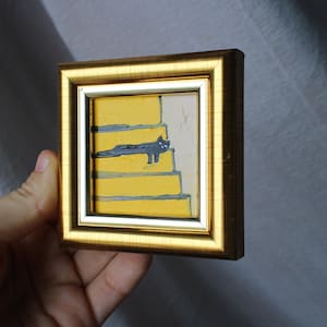 May include: A small framed painting of a black cat sitting on yellow stairs. The painting is in a gold frame.
