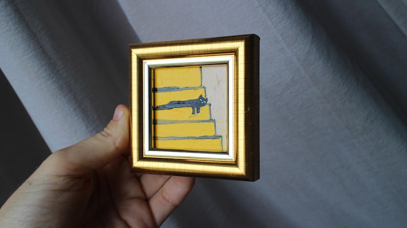 May include: A small framed painting of a black cat sitting on yellow stairs. The painting is in a gold frame.