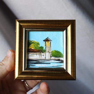 Puede incluir: Una pequeña pintura enmarcada de un edificio con una torre del reloj. El edificio está pintado en tonos de blanco, marrón y verde, con un cielo azul y agua en el fondo.