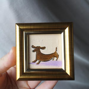 May include: A small, square framed painting featuring a brown dachshund dog. The dog is depicted in a running pose against a textured, off-white background with a purple base. The frame is gold-colored.