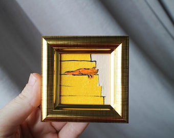May include: A small framed painting of an orange cat lying on yellow stairs. The painting is in a gold frame.