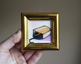 May include: A small framed painting depicting a black and orange box with a black cord attached. The box is resting on a pink background.