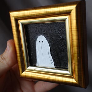 K&ouml;nnte beinhalten: Gerahmtes kleines Kunstwerk mit einer wei&szlig;en Geisterfigur vor einem strukturierten schwarzen Hintergrund. Das Kunstwerk ist in einem goldfarbenen rechteckigen Rahmen eingefasst. Der Geist hat zwei kleine Augenpunkte und eine einfache, flie&szlig;ende Form.