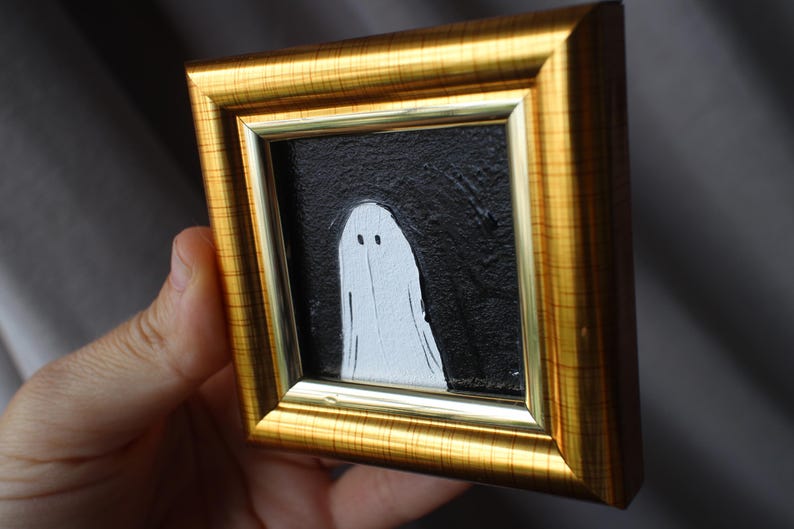 K&ouml;nnte beinhalten: Gerahmtes kleines Kunstwerk mit einer wei&szlig;en Geisterfigur vor einem strukturierten schwarzen Hintergrund. Das Kunstwerk ist in einem goldfarbenen rechteckigen Rahmen eingefasst. Der Geist hat zwei kleine Augenpunkte und eine einfache, flie&szlig;ende Form.