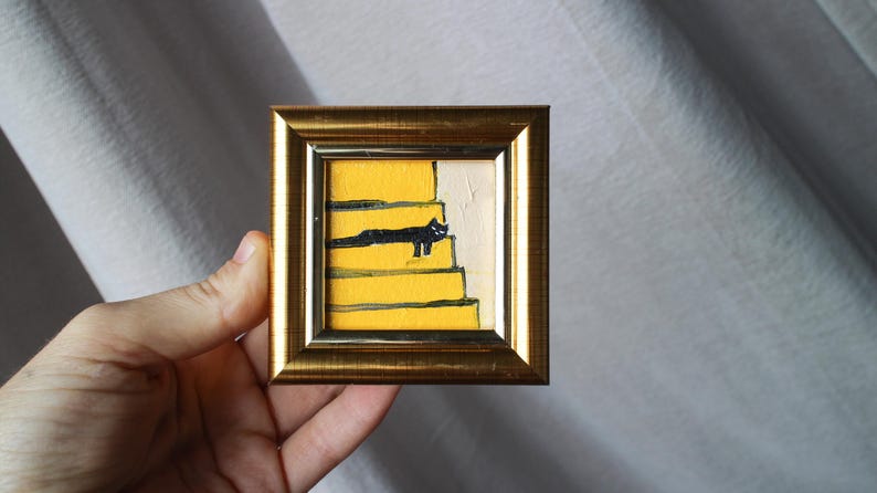 A small framed painting of a black cat sitting on yellow stairs. The painting is in a gold frame.