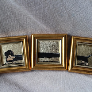 May include: Three small, square paintings in gold frames. Each painting has a gold background with black and beige abstract shapes. The frames are a shiny gold color. The paintings are displayed on a light gray fabric.