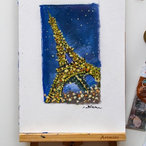 May include: A painting of the Eiffel Tower at night, with a blue background and yellow lights. The painting is on a small easel with the word "Artmate" printed on it.