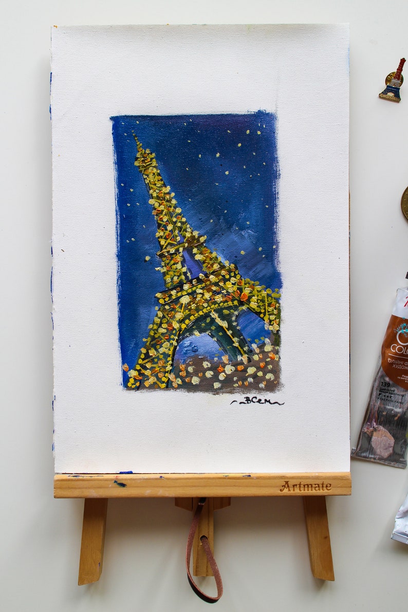 May include: A painting of the Eiffel Tower at night, with a blue background and yellow lights. The painting is on a small easel with the word "Artmate" printed on it.