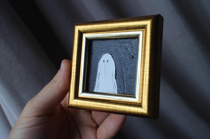 K&ouml;nnte beinhalten: Ein kleines gerahmtes Kunstwerk mit einer wei&szlig;en Geisterfigur vor einem dunkelgrauen Hintergrund. Das Kunstwerk ist in einem goldfarbenen, verzierten Rahmen eingefasst. Der Geist hat ein einfaches, minimalistisches Design.