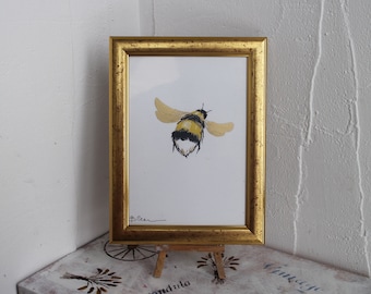 May include: A framed watercolor painting of a bumblebee in flight. The bumblebee is painted in shades of yellow and black, with gold accents. The frame is gold and has a simple design.