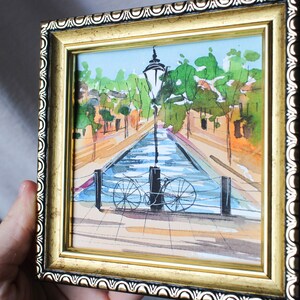 May include: A framed watercolour painting depicting a canal scene with a bicycle resting against a lamppost. The canal is flanked by buildings and trees, and the water is a pale blue colour.