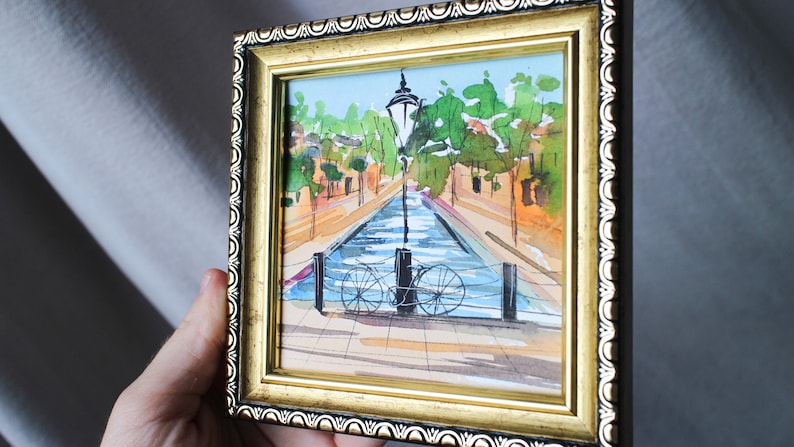 May include: A framed watercolour painting depicting a canal scene with a bicycle resting against a lamppost. The canal is flanked by buildings and trees, and the water is a pale blue colour.