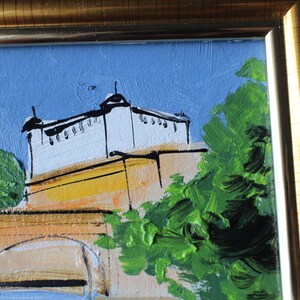 Oil painting of a building with a white facade and black accents, set against a blue sky. The artwork features a gold frame and green foliage. The painting uses thick brushstrokes and a palette of blues, greens, and yellows.