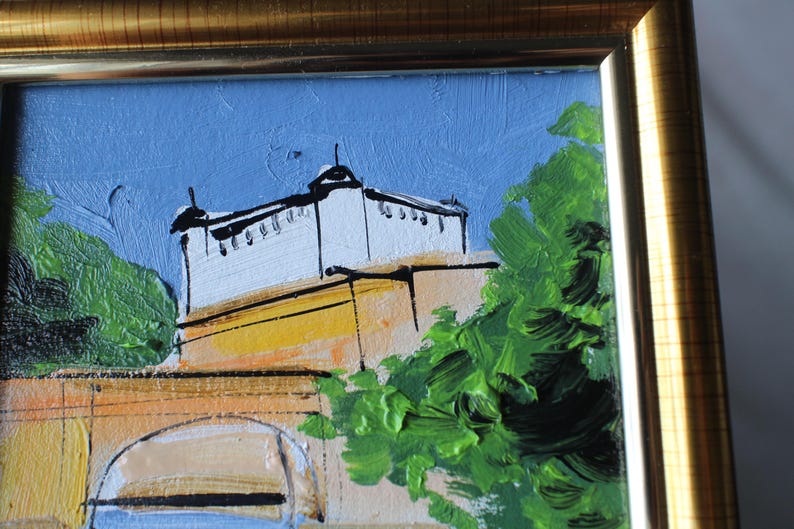 Oil painting of a building with a white facade and black accents, set against a blue sky. The artwork features a gold frame and green foliage. The painting uses thick brushstrokes and a palette of blues, greens, and yellows.