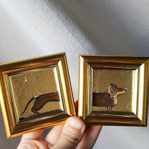 Op de afbeelding: Twee goudkleurige ingelijste afbeeldingen met een zwart-bruin dackel silhouet op een gouden achtergrond.