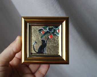May include: A small framed painting of a black cat looking up at a branch with red berries. The background is gold.