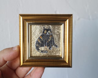May include: A small, square framed artwork featuring a black and orange cat illustration on a gold background. The frame is a warm gold color. The cat has yellow eyes and is sitting. The artwork is held by a hand.