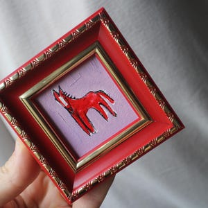 A small, square framed artwork featuring a red horse illustration on a lavender background. The frame is red with gold trim and decorative detailing. The artwork is held in a hand.