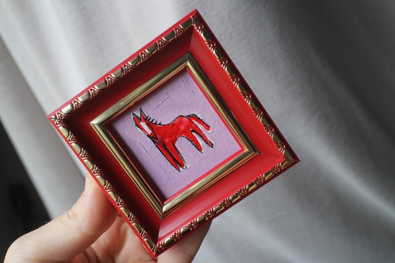 A small, square framed artwork featuring a red horse illustration on a lavender background. The frame is red with gold trim and decorative detailing. The artwork is held in a hand.