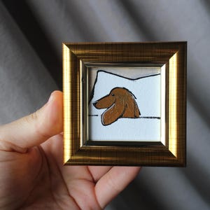 以下が含まれることがあります： 茶色の犬のイラストが描かれた、小さな正方形の額装されたアートワーク。 犬は白い枕の上にいます。 フレームは金色で、アートワークは手で持たれています。