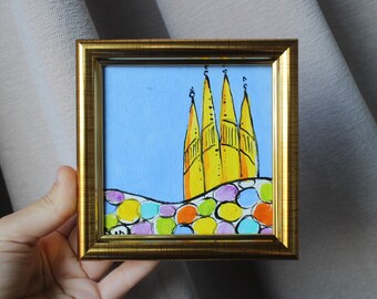 May include: A small, square painting in a gold frame. The artwork depicts a yellow cathedral with four spires against a light blue sky. The base of the cathedral sits atop a colorful, rounded wall with various colored circles.