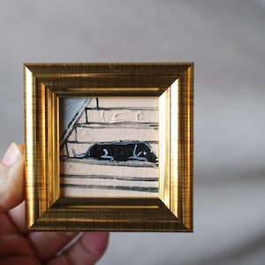 Peut inclure: Une petite peinture encadrée d'un chien noir couché sur un escalier. La peinture est dans un cadre doré et a un fond blanc.