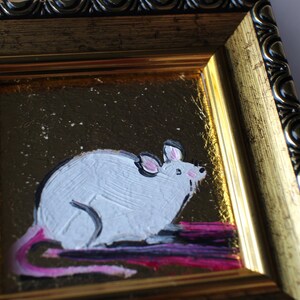 May include: A white mouse with a pink tail is painted on a gold background with a sparkly texture. The painting is framed in a gold and black ornate frame.
