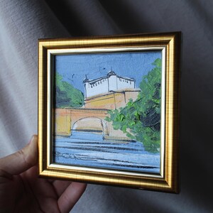 Small framed painting with a gold-colored frame. The artwork depicts a bridge over water with a building in the background. The painting uses blue, yellow, green, and white colors. The artwork is held by a hand.