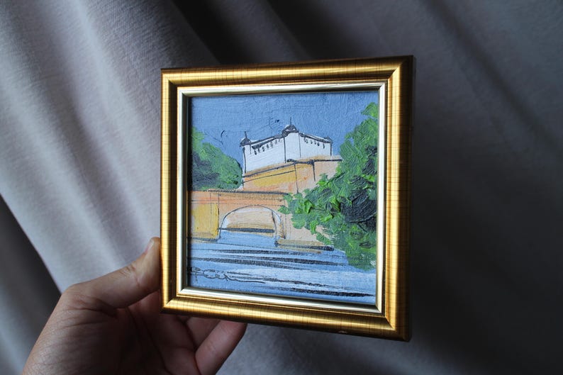 Small framed painting with a gold-colored frame. The artwork depicts a bridge over water with a building in the background. The painting uses blue, yellow, green, and white colors. The artwork is held by a hand.